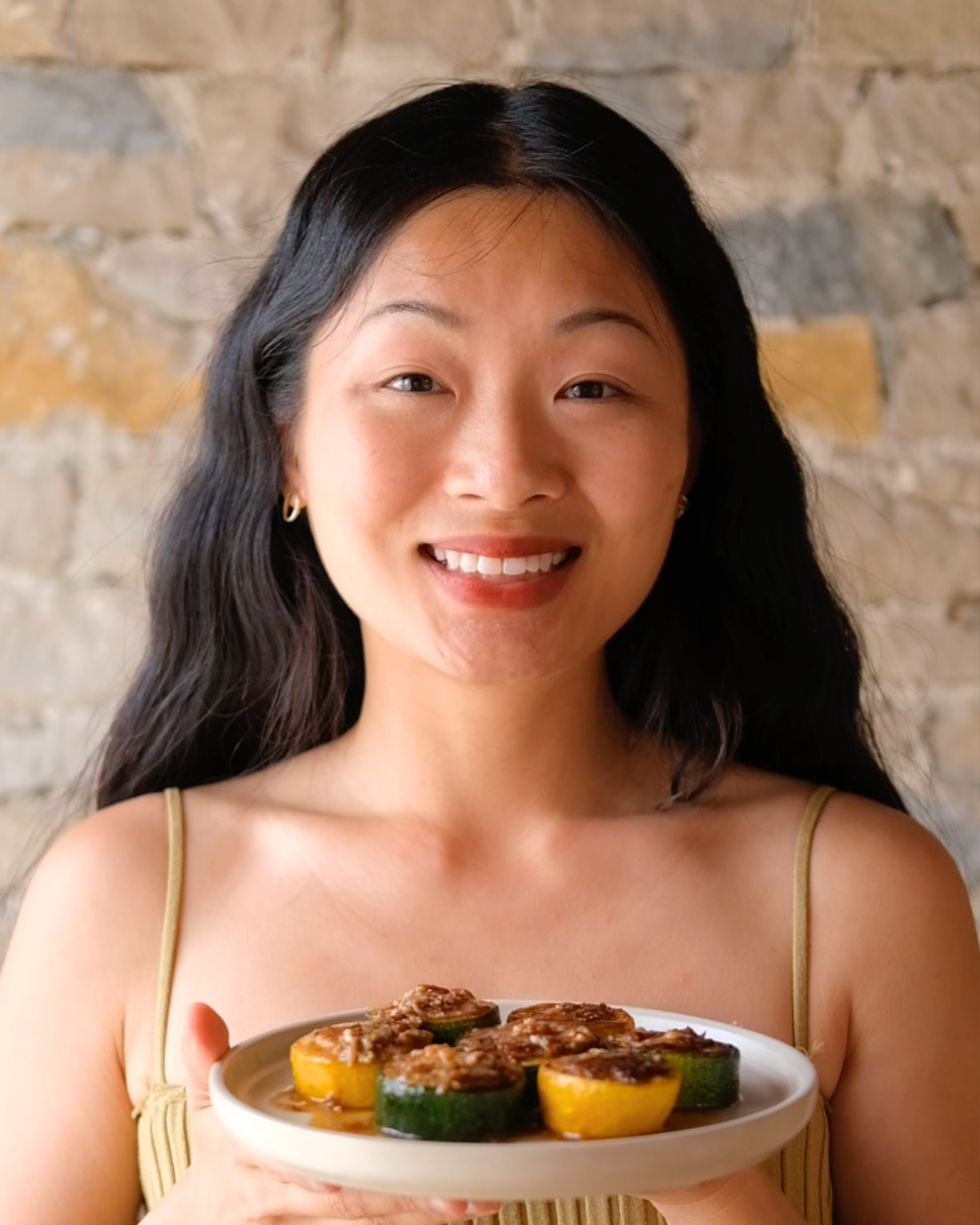 Woman with long dark wavy hair smiling and holding a plate of caramelised zucchini rounds topped with savoury sauce, standing in front of a stone wall.